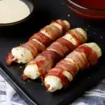 bacon wrapped in cheese on a black plate with dipping sauce next to the bowl full of ketchup